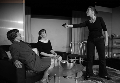 Bild zeigt einen Ausschnitt aus dem Theaterstück 'Kaltgestellt'. Man sieht eine Frau, die in einem Wohnzimmer steht, eine Waffe in der Hand hält und diese auf zwei Frauen richtet